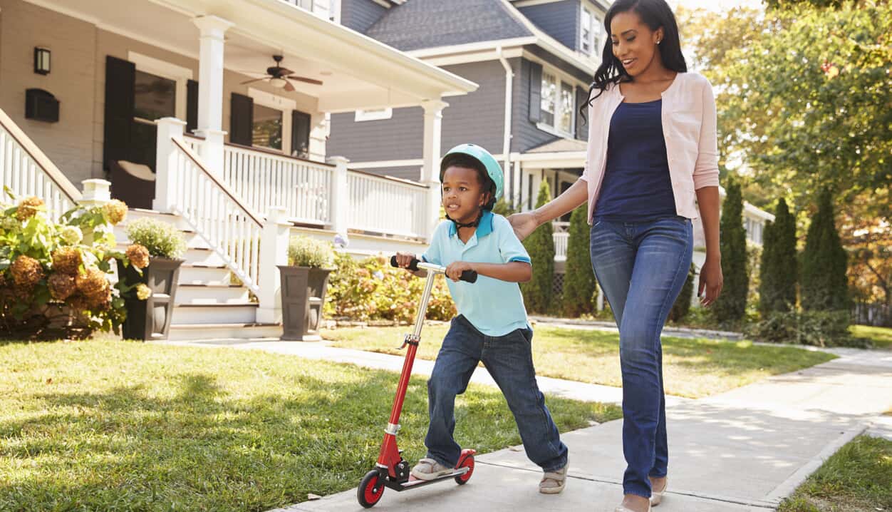 A mother walks with her son riding a scooter on a suburban sidewalk